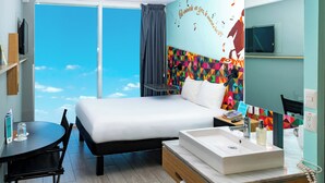 Habitación de hotel con cama, escritorio, baño con lavamanos y un amplio ventanal con vistas al cielo.