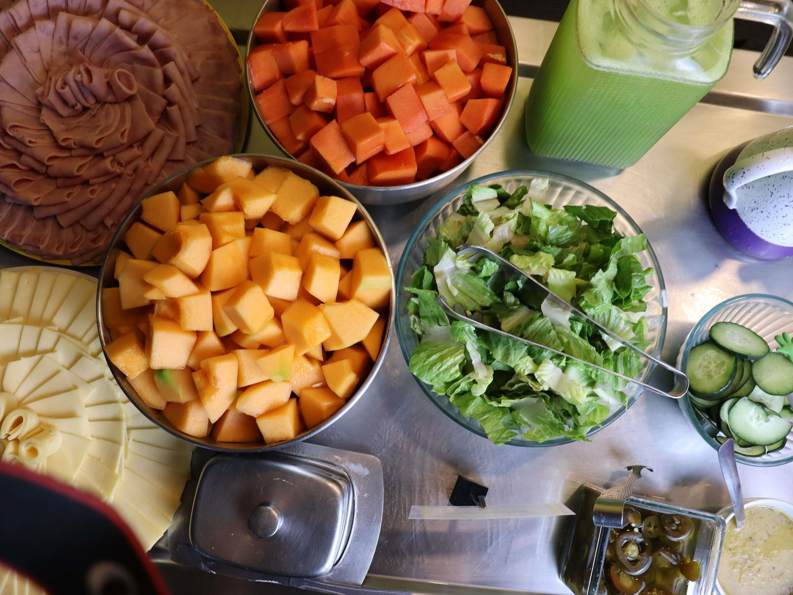 Una mesa de buffet con diversos alimentos, incluyendo carnes en rodajas, melón en cubos, un batido verde, una ensalada y pepinos en rodajas.