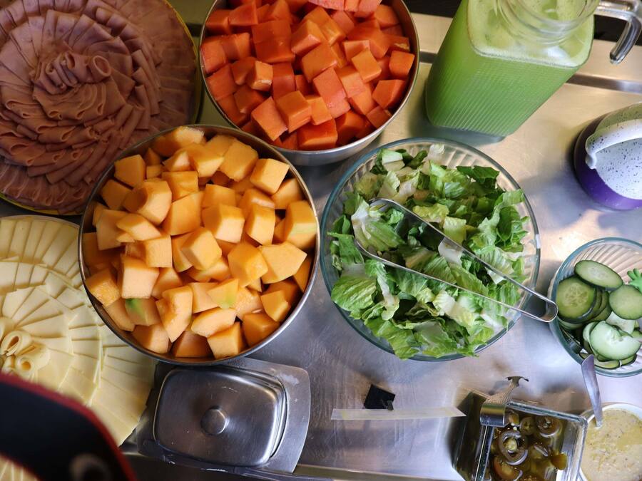 Una mesa de buffet con diversos alimentos, incluyendo carnes en rodajas, melón en cubos, un batido verde, una ensalada y pepinos en rodajas.