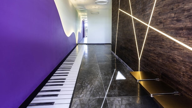A modern hallway with a piano, a wooden wall with geometric lighting, and a purple wall. - ibis Styles Merida Galerias (Mérida)