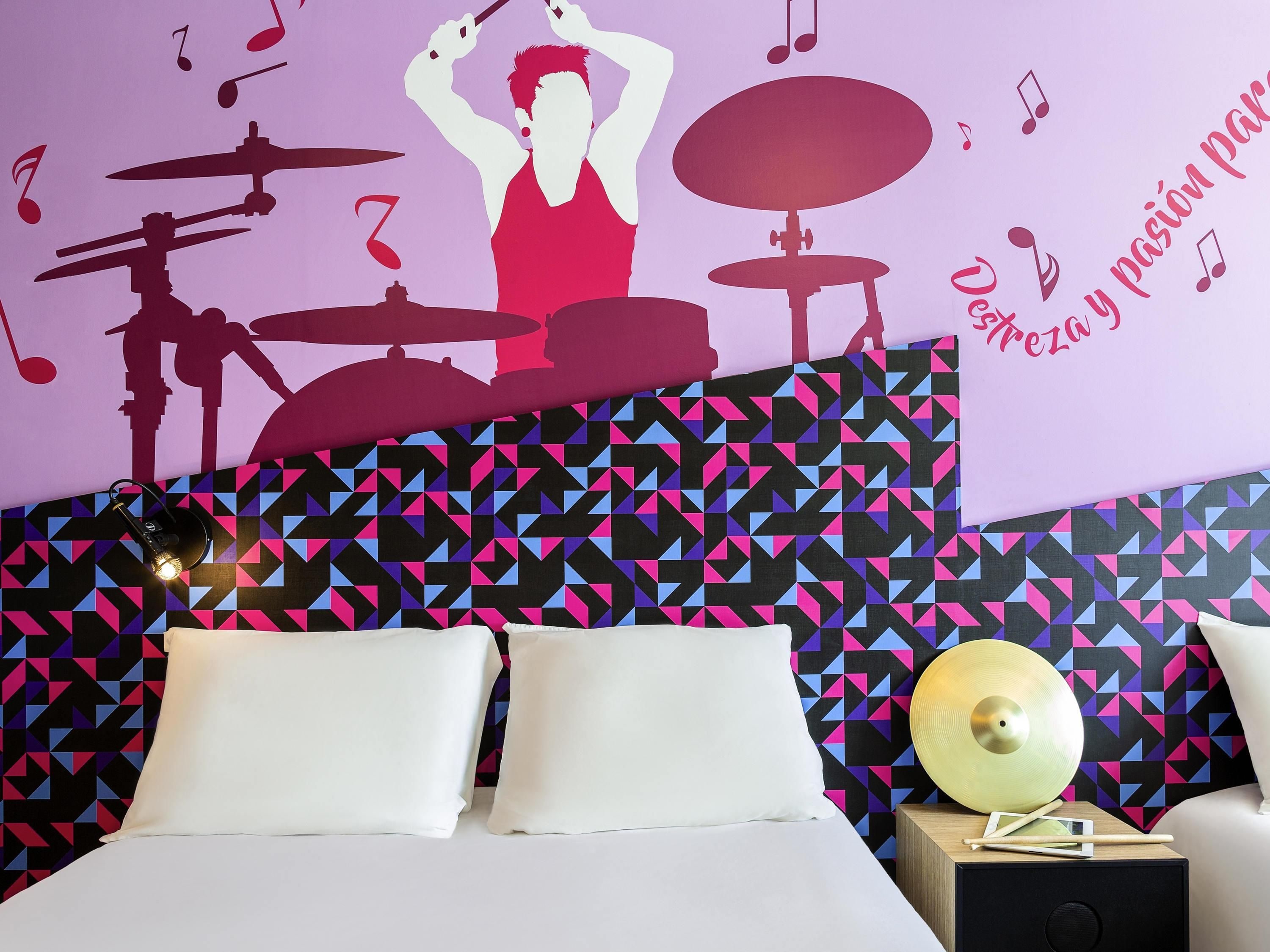 A hotel room with a bed, pillows, a nightstand with a lamp, a decorative wall with geometric patterns, and a wall decal of a drummer.
