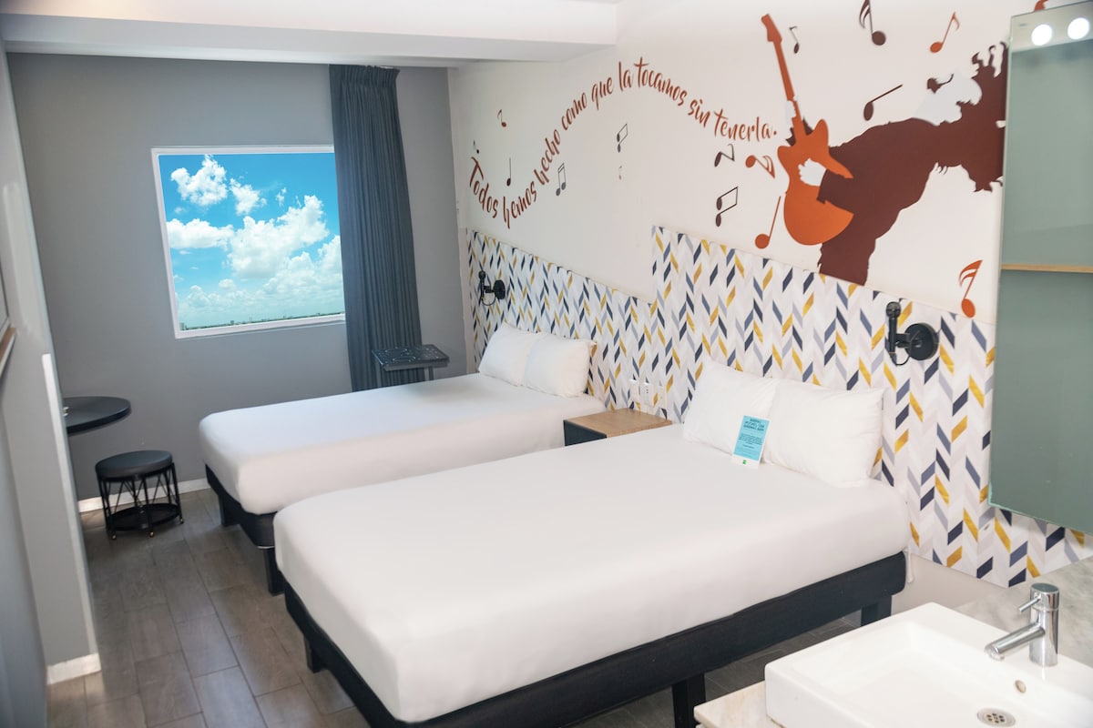A hotel room with two beds, a window with a view, a wall with a musical theme, and a bathroom with a mirror and sink.