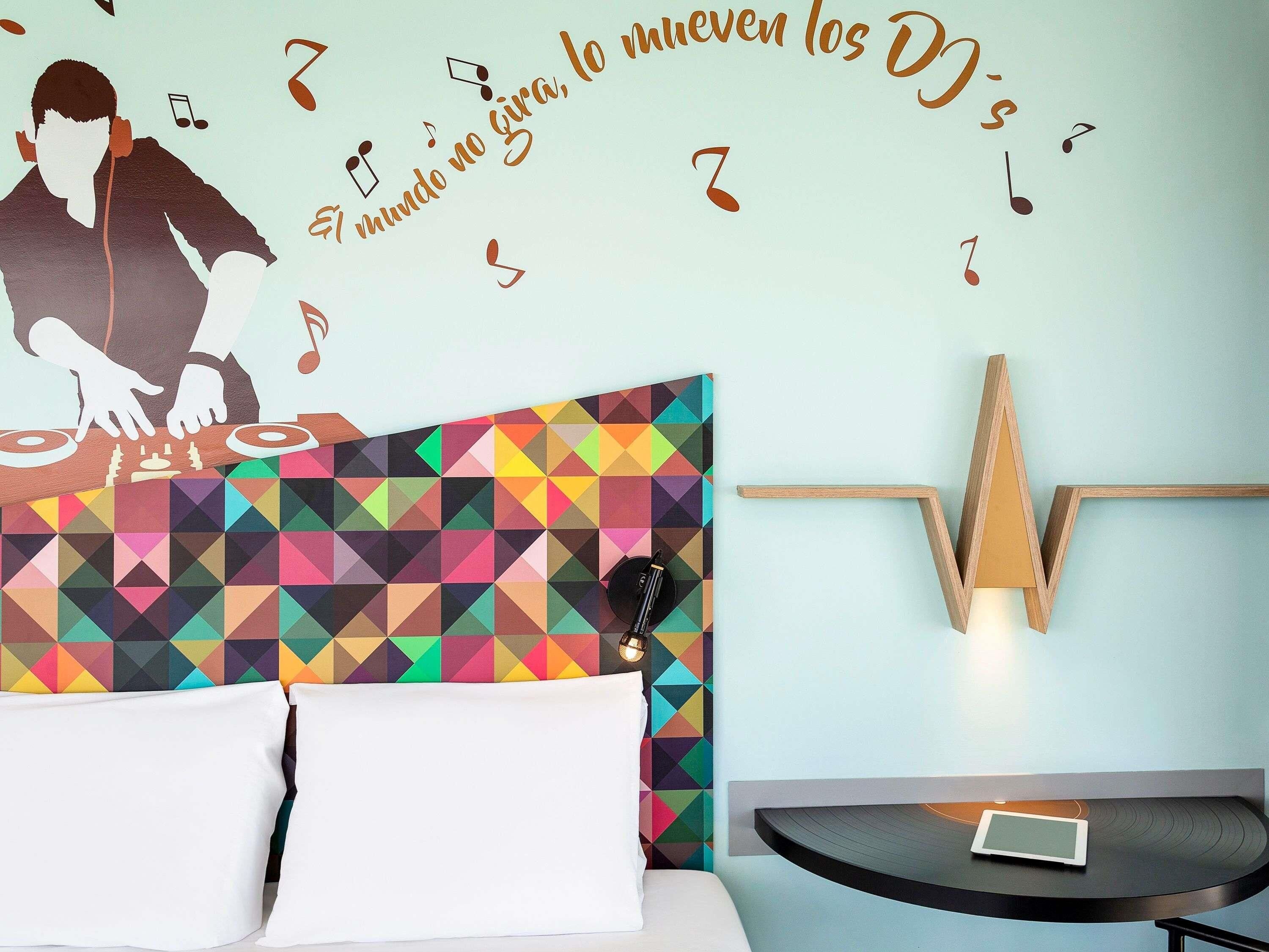 Une chambre d’hôtel avec une tête de lit aux motifs géométriques colorés, un lit avec du linge de lit blanc, une table de chevet avec une tablette et un luminaire fixé au mur.