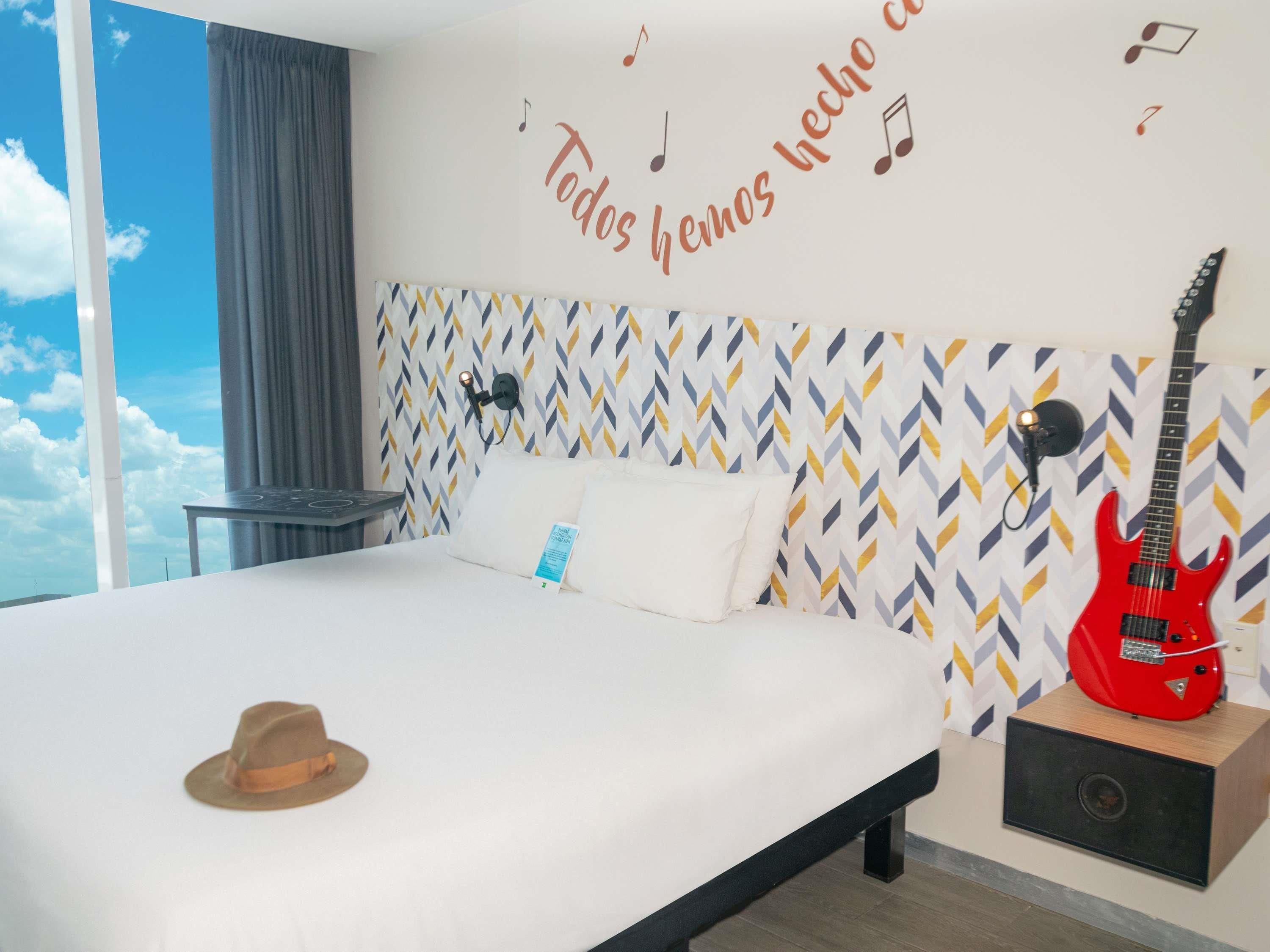 Una habitación de hotel con una cama, una guitarra eléctrica roja, un sombrero y una pared con un diseño de notas musicales.