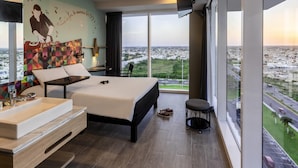 A modern hotel room with a large bed, a bathroom with a sink and mirror, a small stool, and a city view. - ibis Styles Merida Galerias (Mérida)