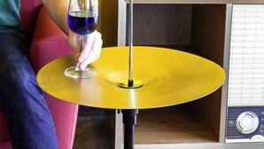 Eine Person sitzt auf einem roten Stuhl vor einem gelben Tisch und hält ein Glas mit blauer Flüssigkeit. Eine rote elektrische Gitarre ist an einer gelben Wand angebracht.