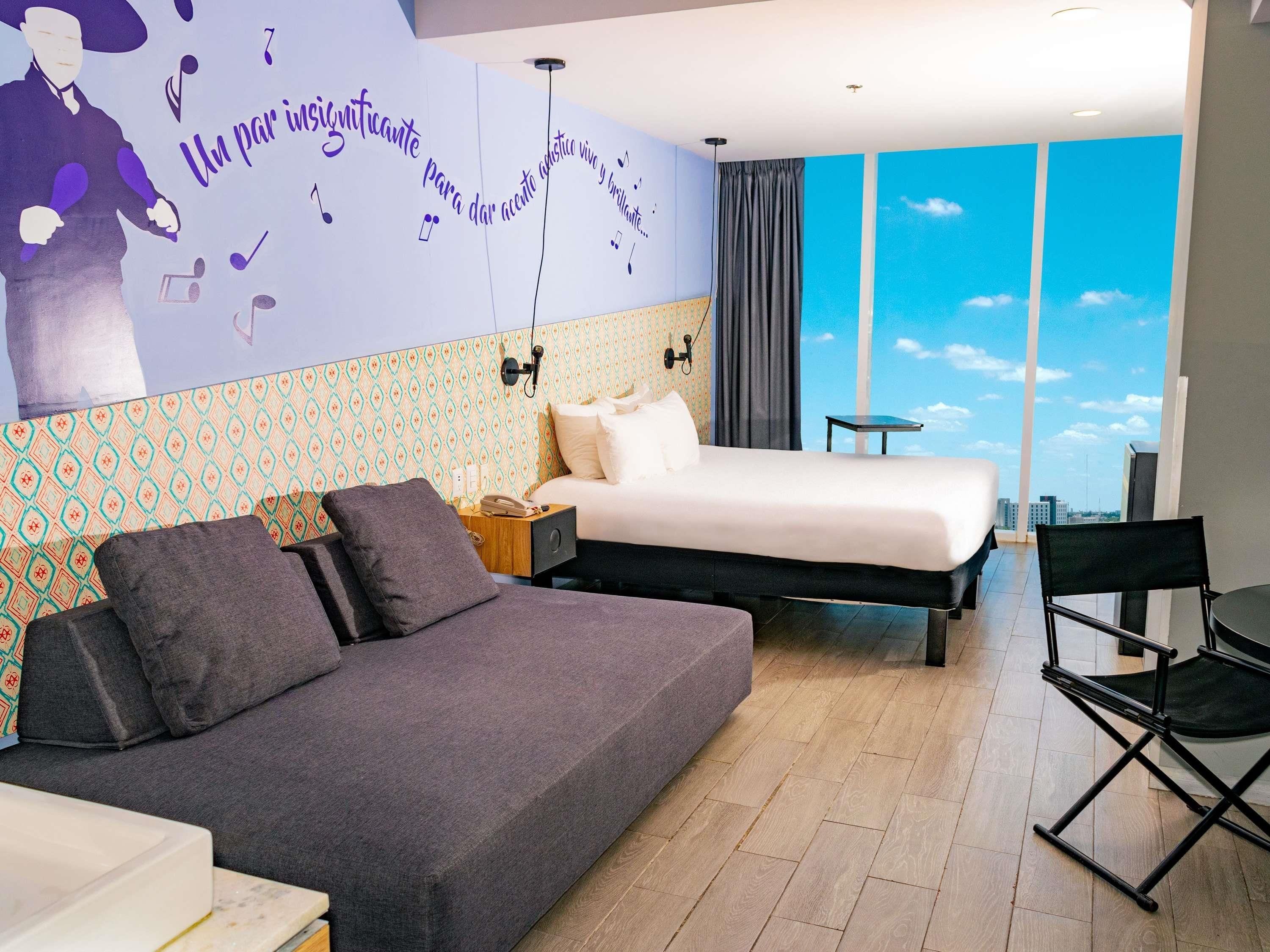 A modern hotel room with a large bed, a sofa, a desk, and a view of the sky through large windows.