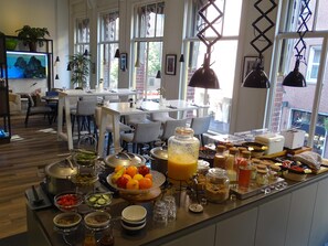 Ein Frühstücksbuffet mit verschiedenen Speisen und Getränken, einem Fernseher und einer Sitzecke mit Tischen und Stühlen.