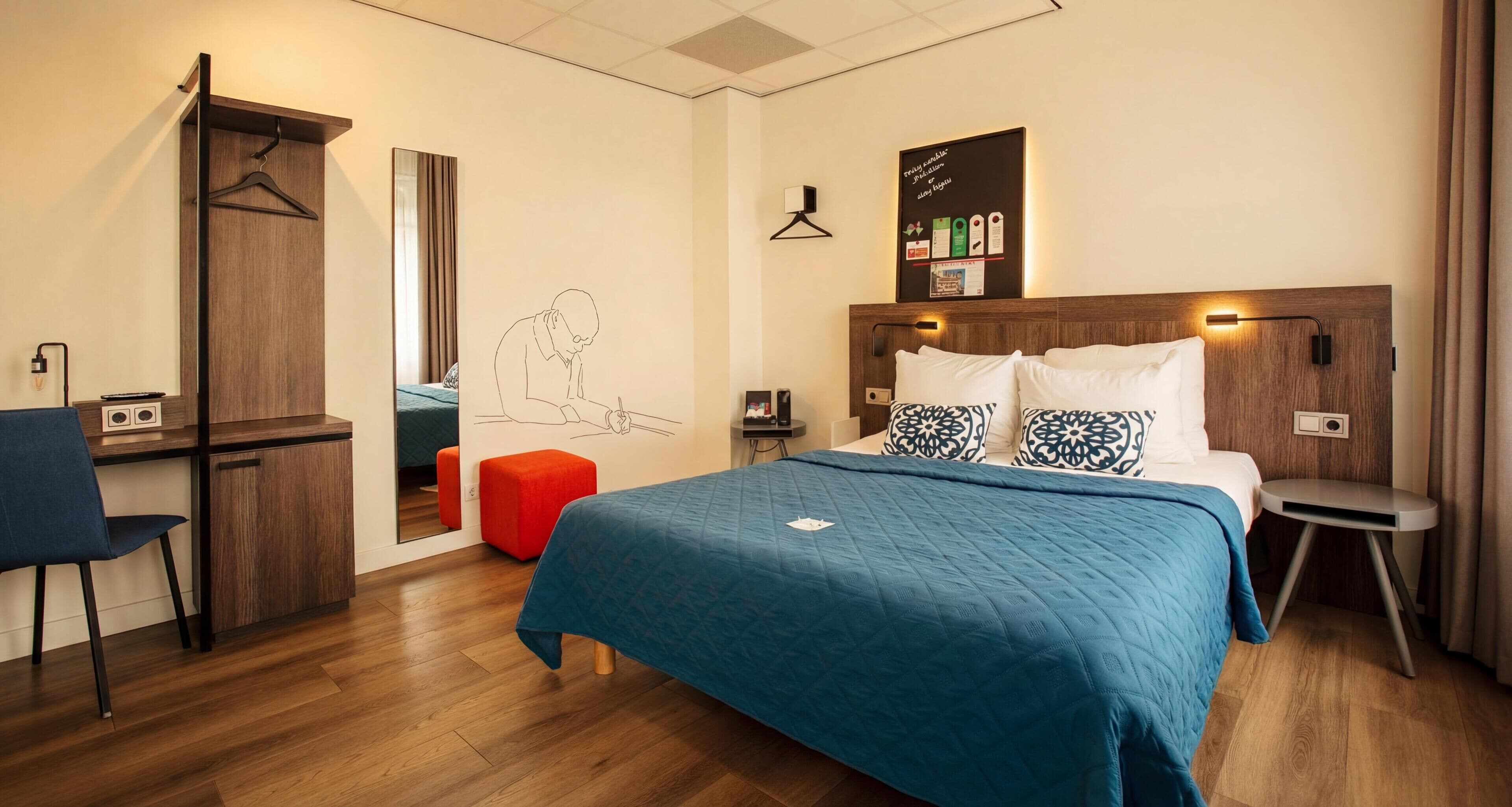 A hotel room with a bed, a desk, a chair, a wardrobe, and a wall with a hanging light fixture and a framed picture.