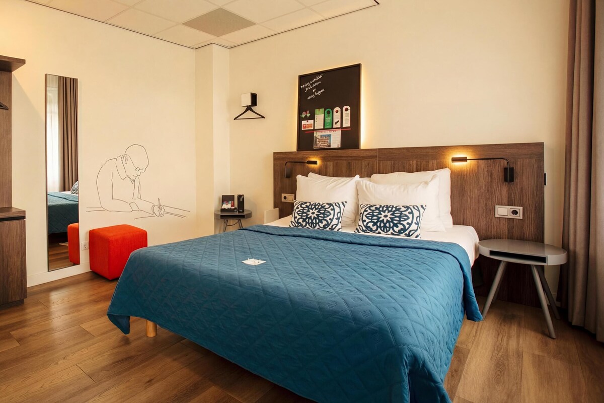 Habitación de hotel con cama, escritorio, silla, armario y una pared con lámpara colgante y un cuadro enmarcado.