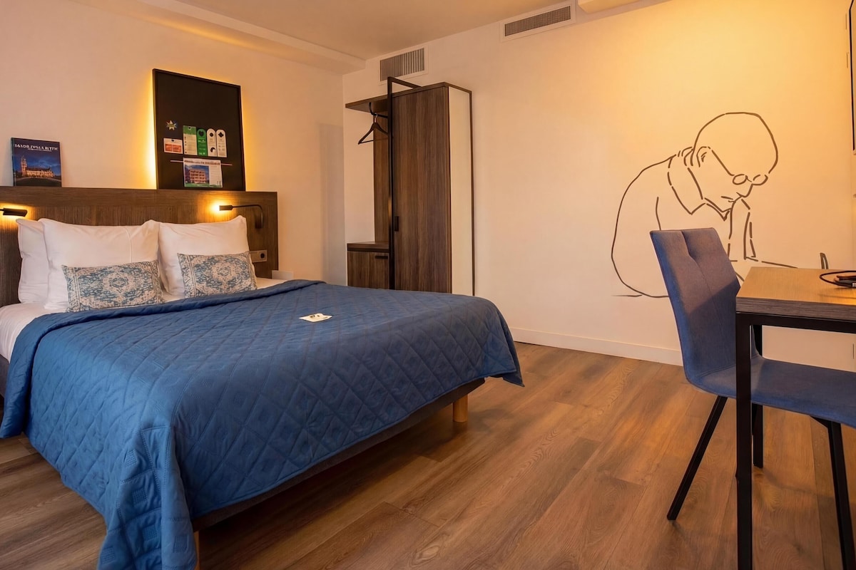 Habitación de hotel con cama, mesita de noche y escritorio con silla. Se observa una estantería de pared con libros y una obra de arte en la pared que representa a una persona.