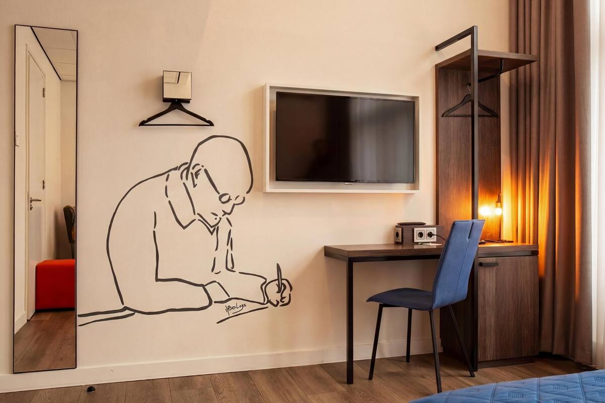 Habitación de hotel con escritorio, silla, televisor y espejo montado en la pared.