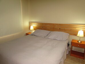 Ein ordentlich gemachtes Bett mit einem Holzkopfende, zwei Nachttische mit Lampen und einer schlichten Wand.