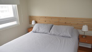 Een net opgemaakt bed met een houten hoofdbord, twee nachtkastjes met lampen, en een raam dat natuurlijk licht binnenlaat.
