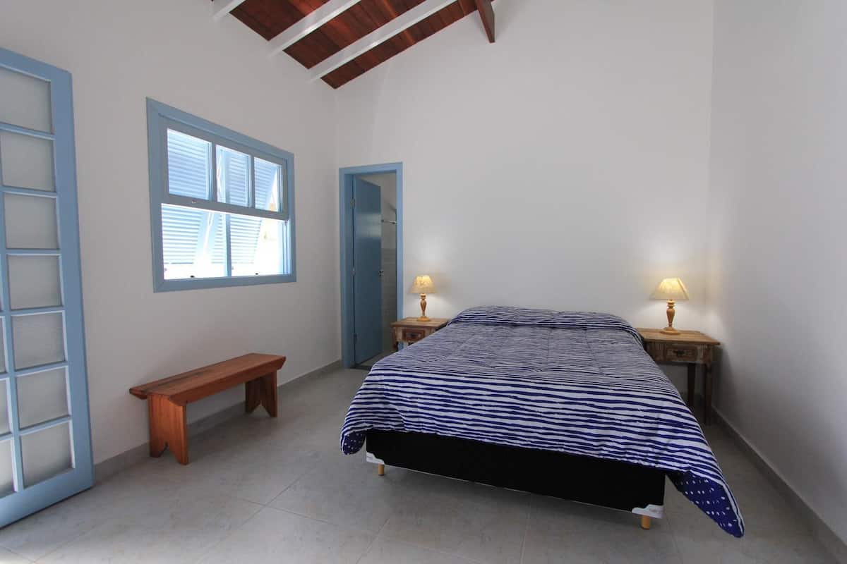 Un dormitorio con una cama, dos mesitas de noche con lámparas, un banco de madera, una ventana con persianas y una puerta.