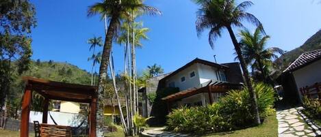 A garden with a wooden pergola, palm trees, a stone pathway, and a house with a tiled roof.