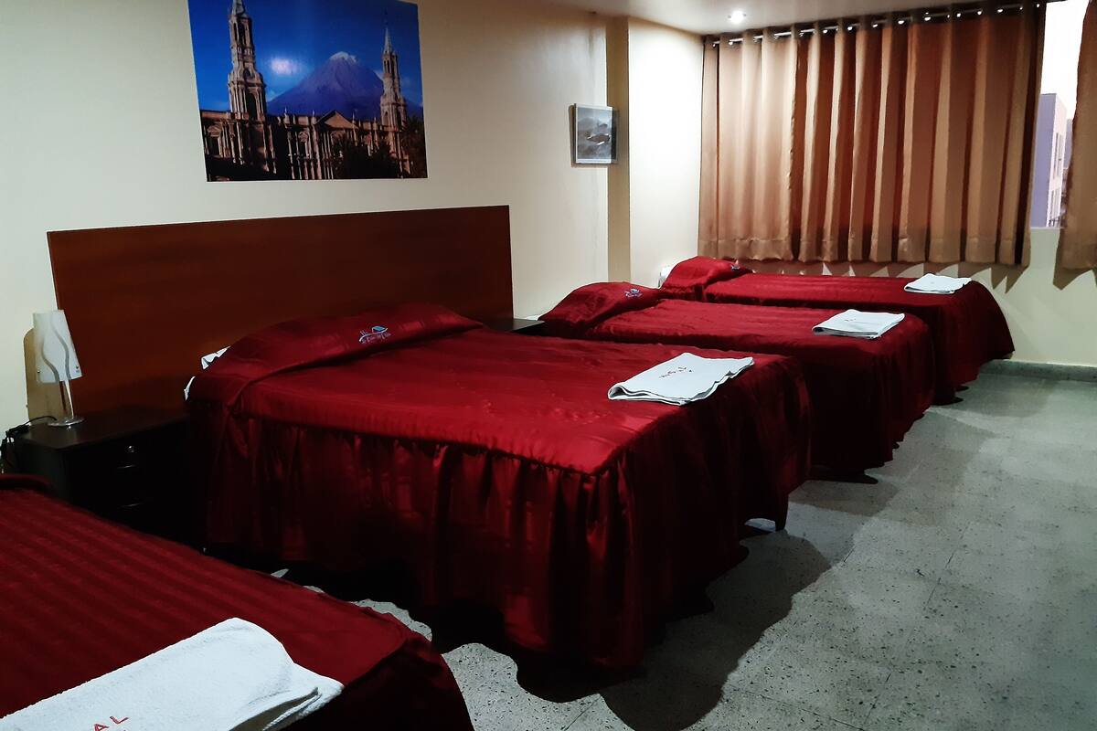 A hotel room with four beds, each covered with red bedding. There is a painting of a church on the wall, a window with curtains, and a small table with a lamp.
