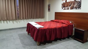 Quarto de hotel com cama, mesa de cabeceira, janela com cortinas, quadro na parede e chão ladrilhado.
