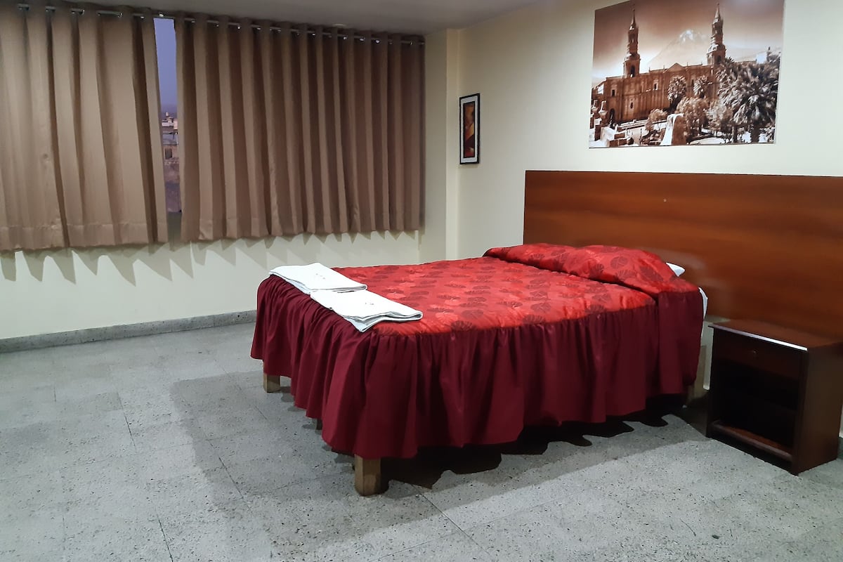 A hotel room with a bed, a nightstand, a window with curtains, a wall-mounted picture, and a tiled floor.