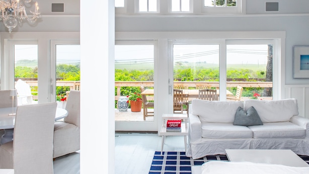 Ein großzügiges Wohnzimmer mit einem weißen Sofa, einem Esstisch und einer Chandelier. Große Fenster bieten Blick auf Grünflächen und eine Holzterrasse draußen.
