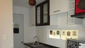 A compact kitchen with a dark countertop, a sink, a stove, and a refrigerator. There are cabinets above the counter and a door leading to another room. - 36.- HNA Paseo Peñuelas (La Serena)