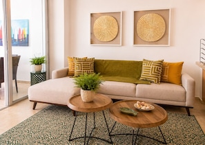 A living room with a sofa, two round wooden tables, a green patterned rug, and two wall-mounted decorative pieces.