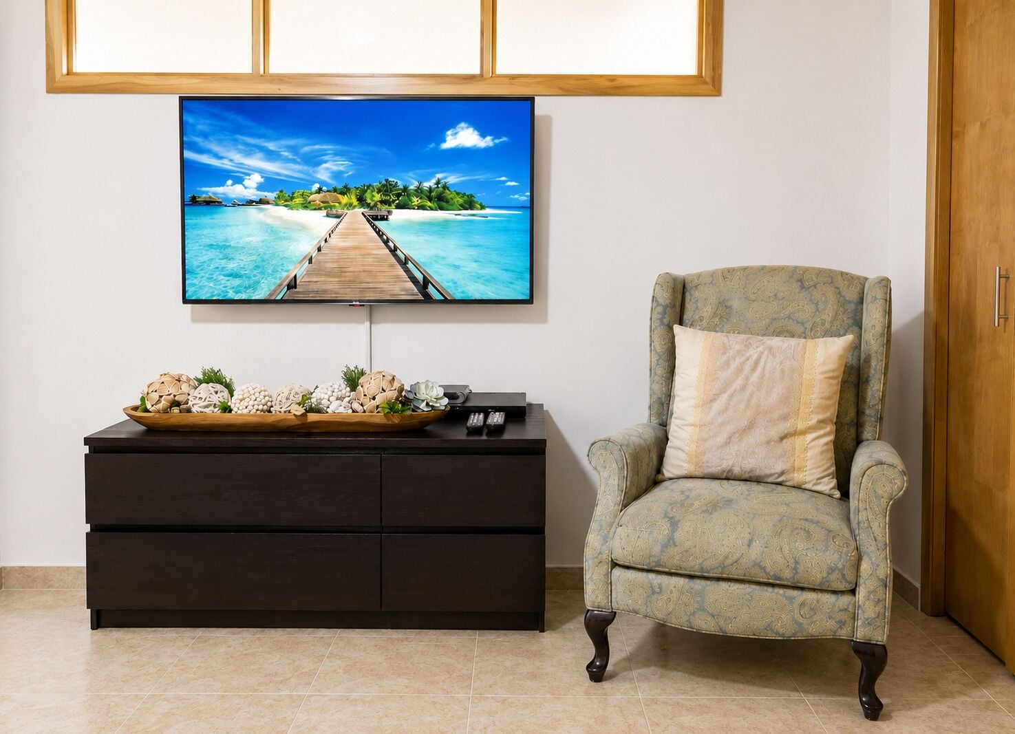Ein Zimmer mit Fernseher, der eine tropische Strandansicht zeigt, einem dunklen Holzschrank und einem gemusterten Sessel.