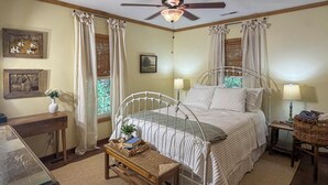Un dormitorio con cama, lámparas de noche, mesitas de noche, una ventana con cortinas, un cuadro en la pared y un ventilador de techo.