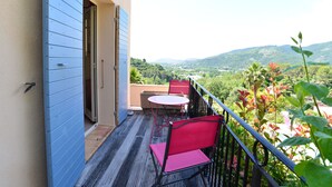 A balcony with a table and chairs, a view of a green landscape, and a building with blue siding.