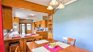 A dining area with a wooden table, chairs, and a bowl of fruit. There is a kitchen in the background with wooden cabinets and a window.