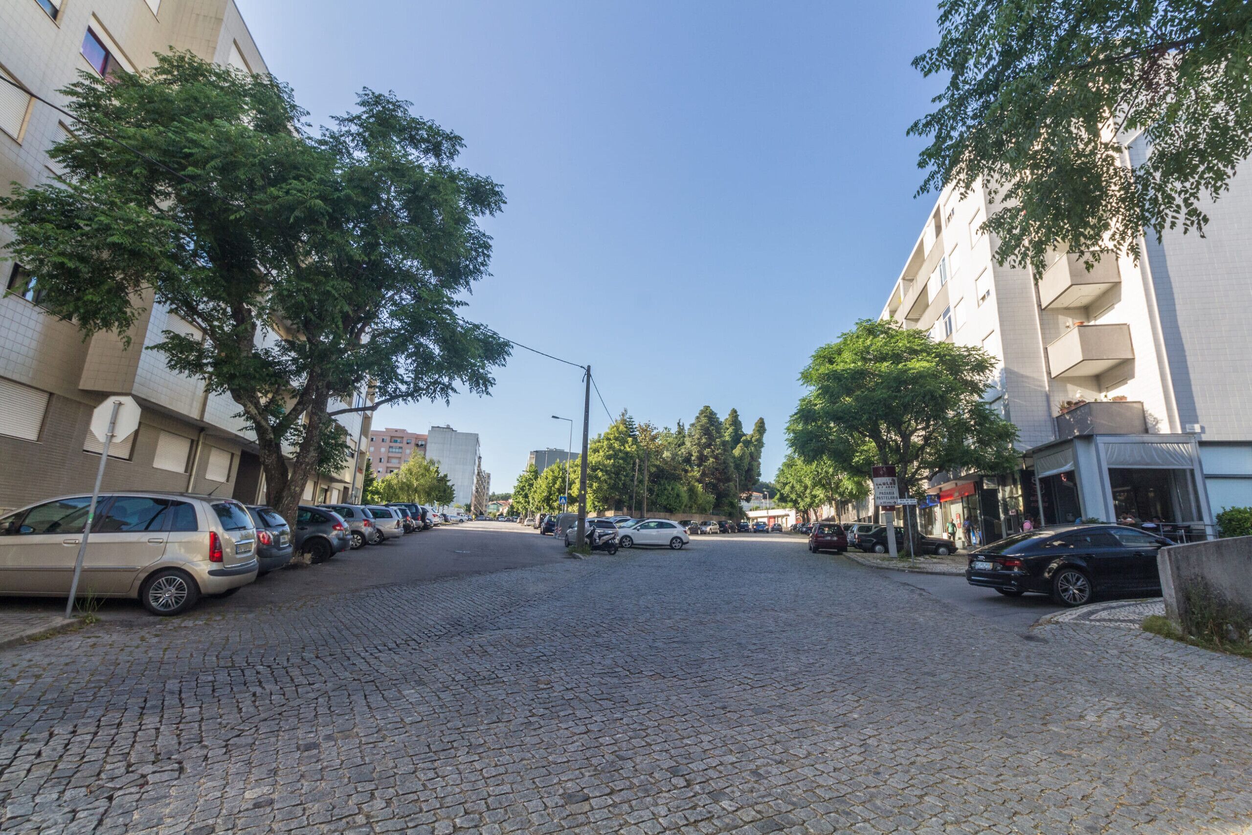 Une rue pavée bordée de voitures garées et d’arbres, qui mène à un immeuble doté de balcons.