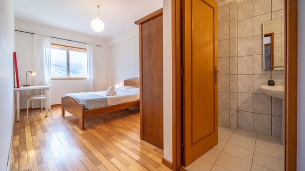 Une chambre à coucher avec un lit en bois, un bureau, une chaise, une fenêtre donnant sur un beau paysage, et une salle de bain avec un lavabo et un miroir.