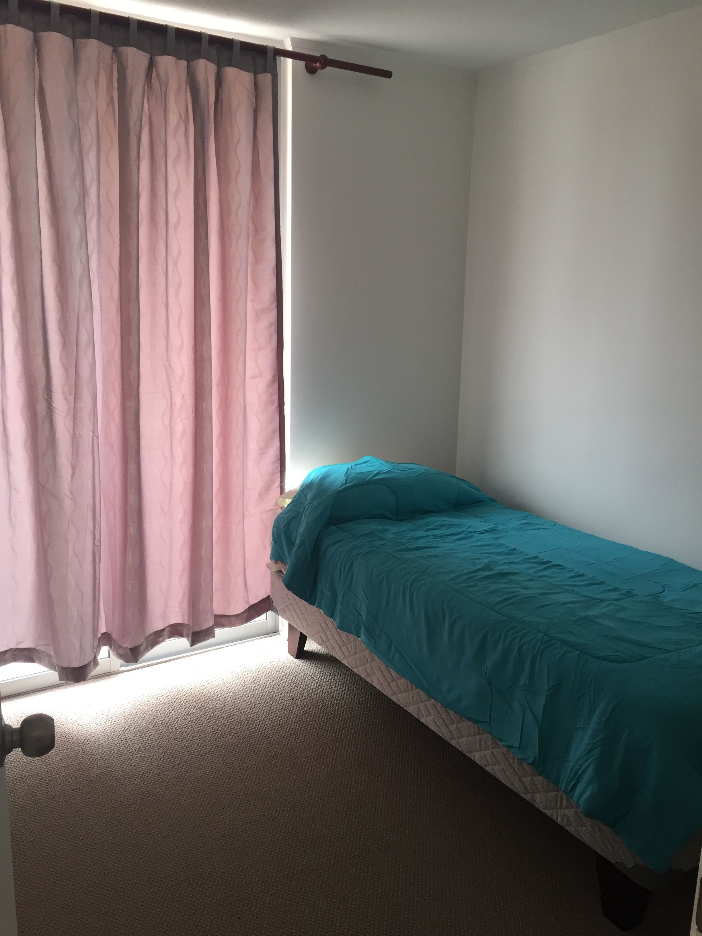Un lit simple avec une couvre-lit turquoise dans une pièce dotée d’une fenêtre ornée d’un rideau rose.