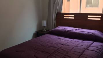 Una cama con colcha morada, cabecera de madera, mesita de noche con lámpara y ventana con cortinas.