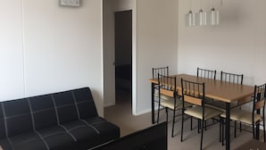 Un salon moderne avec un canapé noir, une table à manger en bois avec des chaises, et une petite œuvre d’art blanche accrochée au mur.