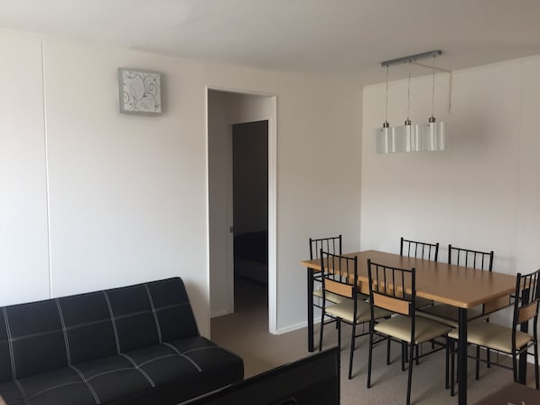 A modern living room with a black sofa, a wooden dining table with chairs, and a small white artwork on the wall.