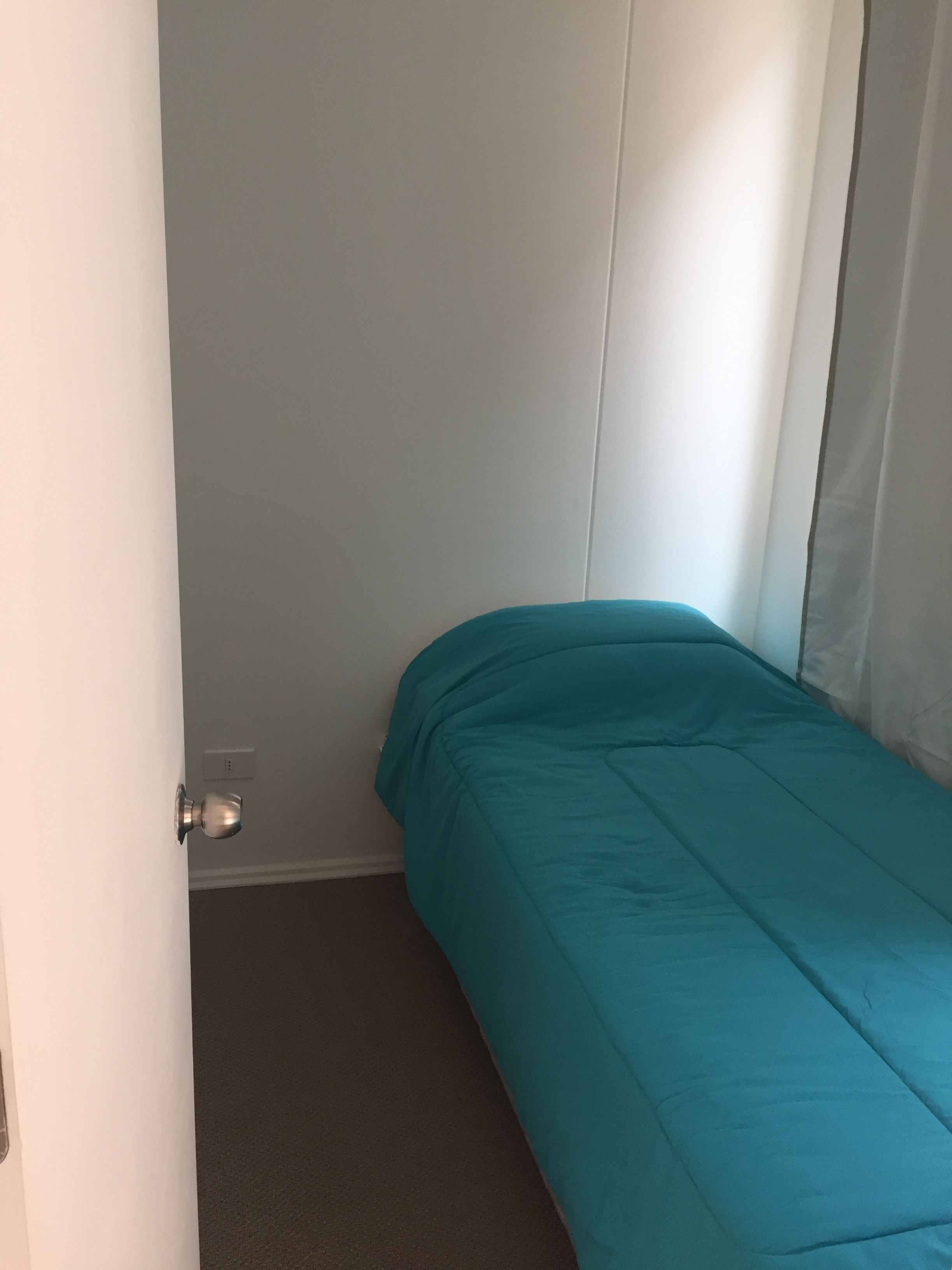 A small, single-bed room with a teal bedspread, white walls, and a door with a silver handle.