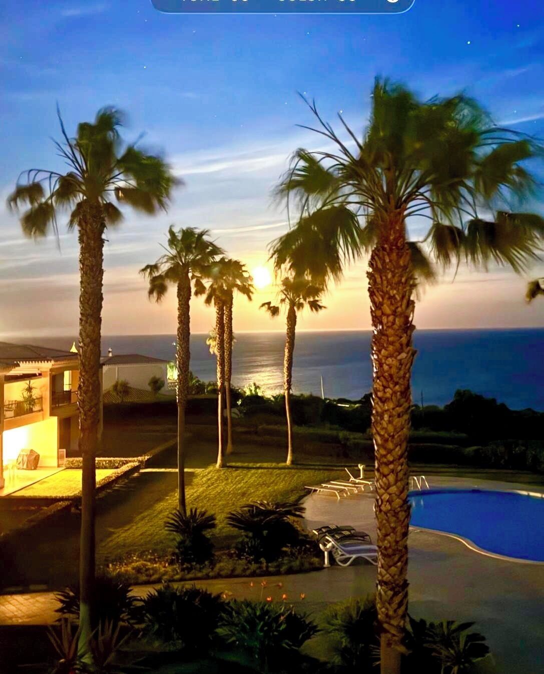 Área da piscina com palmeiras, um edifício e vista para o oceano ao pôr do sol.