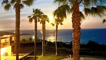 Área da piscina com palmeiras, um edifício e vista para o oceano ao pôr do sol.
