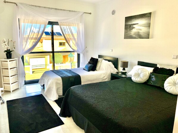 A hotel room with two beds, a window with sheer curtains, a small table with a lamp, and a wall-mounted picture.