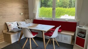 Un cuarto acogedor con pared de madera, una ventana con vista a follaje, una mesa blanca con sillas y un banco rojo.