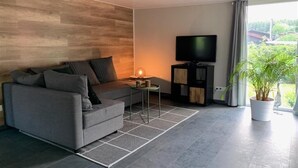 Una sala moderna con un sofá gris, una mesita auxiliar, un televisor de pantalla plana sobre un soporte negro, una planta en maceta y vistas a espacios verdes al exterior.