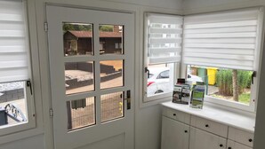Una habitación con una puerta blanca, una ventana con persianas, un mueble pequeño y vistas al exterior.