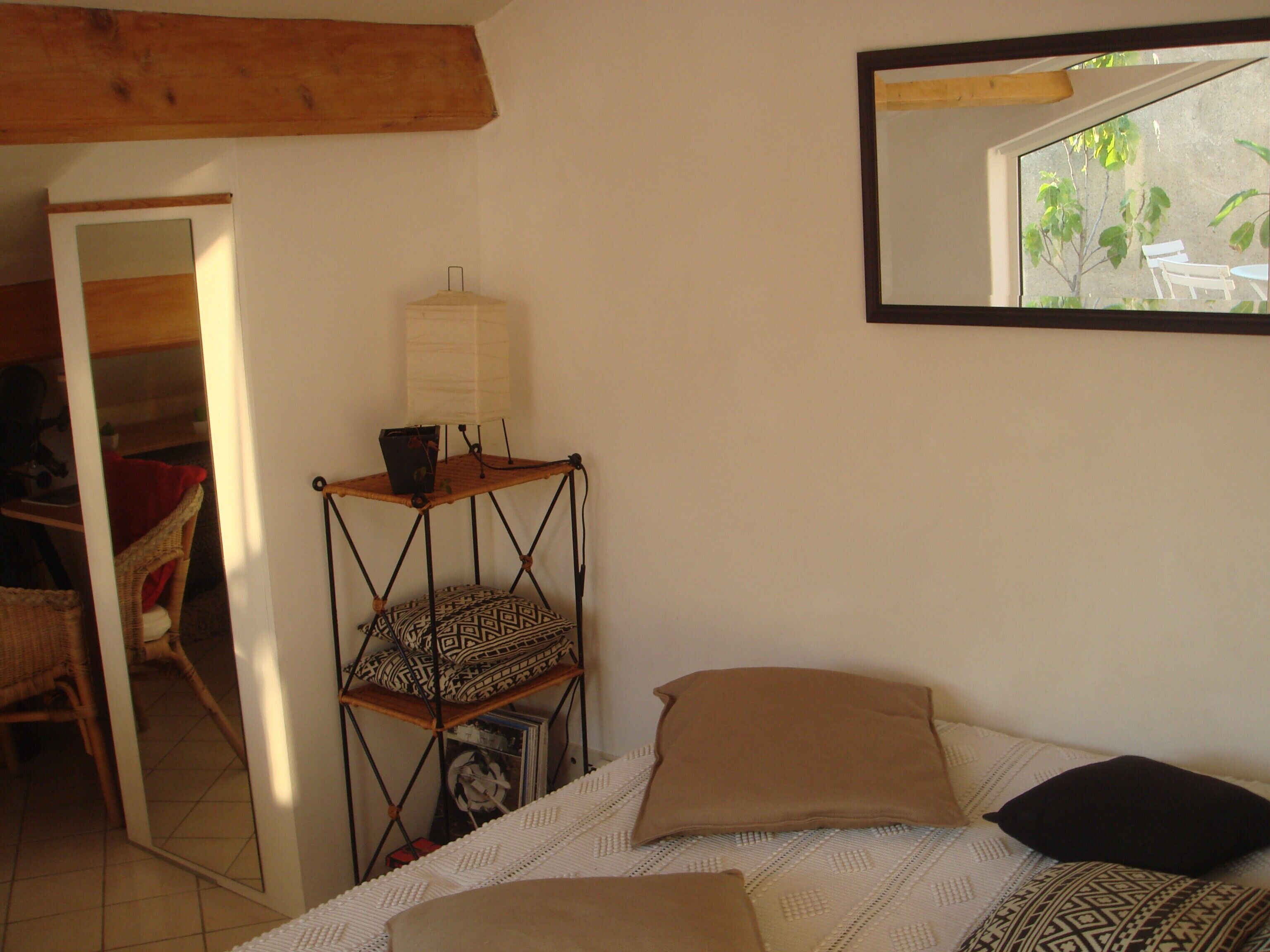 A bedroom with a bed, pillows, a small wooden shelf with a lamp, a mirror, and a view of the outdoors through a window.