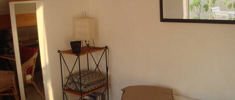 A bedroom with a bed, pillows, a small wooden shelf with a lamp, a mirror, and a view of the outdoors through a window.
