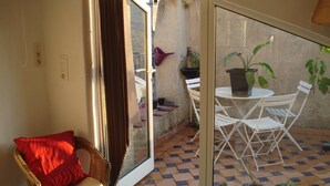 Una habitación con una silla de mimbre, un cojín rojo, una puerta de cristal que da a un espacio exterior con suelo de baldosas, una mesa blanca con sillas y plantas en macetas.