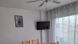 Un comedor con una mesa y sillas, un ventilador de techo, un televisor en la pared y una ventana con cortinas.