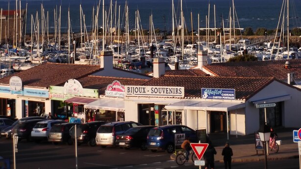 Un puerto deportivo con numerosos barcos amarrados, una tienda de recuerdos y algunos coches estacionados al frente.