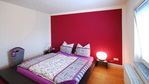 Ein Schlafzimmer mit roter Akzentwand, einem Bett mit gemusterten Betttextilien, einer Nachttischlampe und einem Heizgerät.
