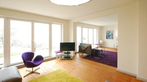 Ein geräumiges Wohnzimmer mit großen Fenstern, einem Flachbildfernseher, einem violetten Sessel und Holzboden.